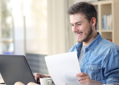 Homem sorrindo olhando computador e segurando papel